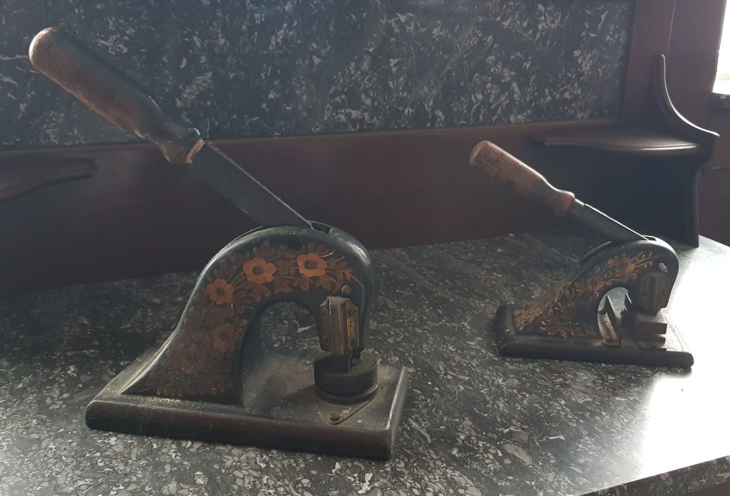 two metal stamp that look like old fashioned hole punches stood on a marble table. Paper is put under the stamp and an arm pushed down to indent the lettering onto the paper.