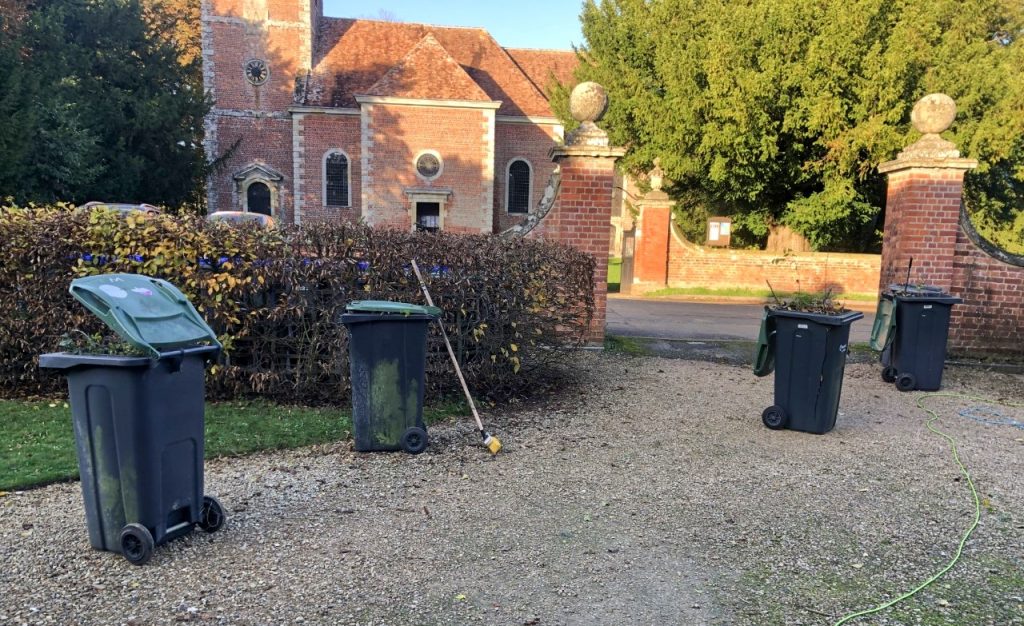 wheelie bins full of hedge clippings in front of a trimmed beech hedge. There is a red brick church in the background